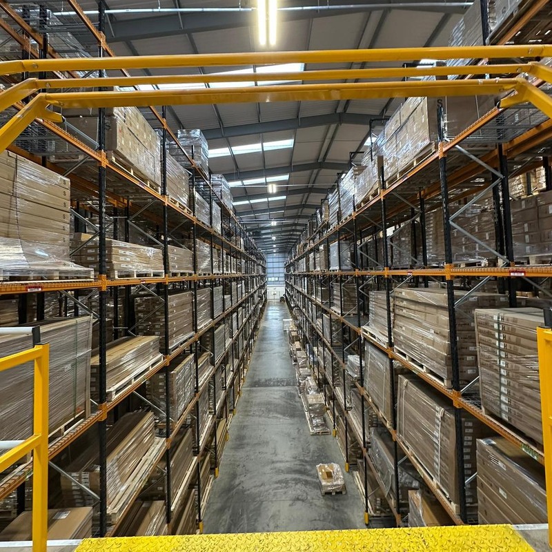 A view of a warehouse with each side of filled with shelving covered in cardboard boxes. The view is framed by the yellow bars of a cherry picker viewing platform.
